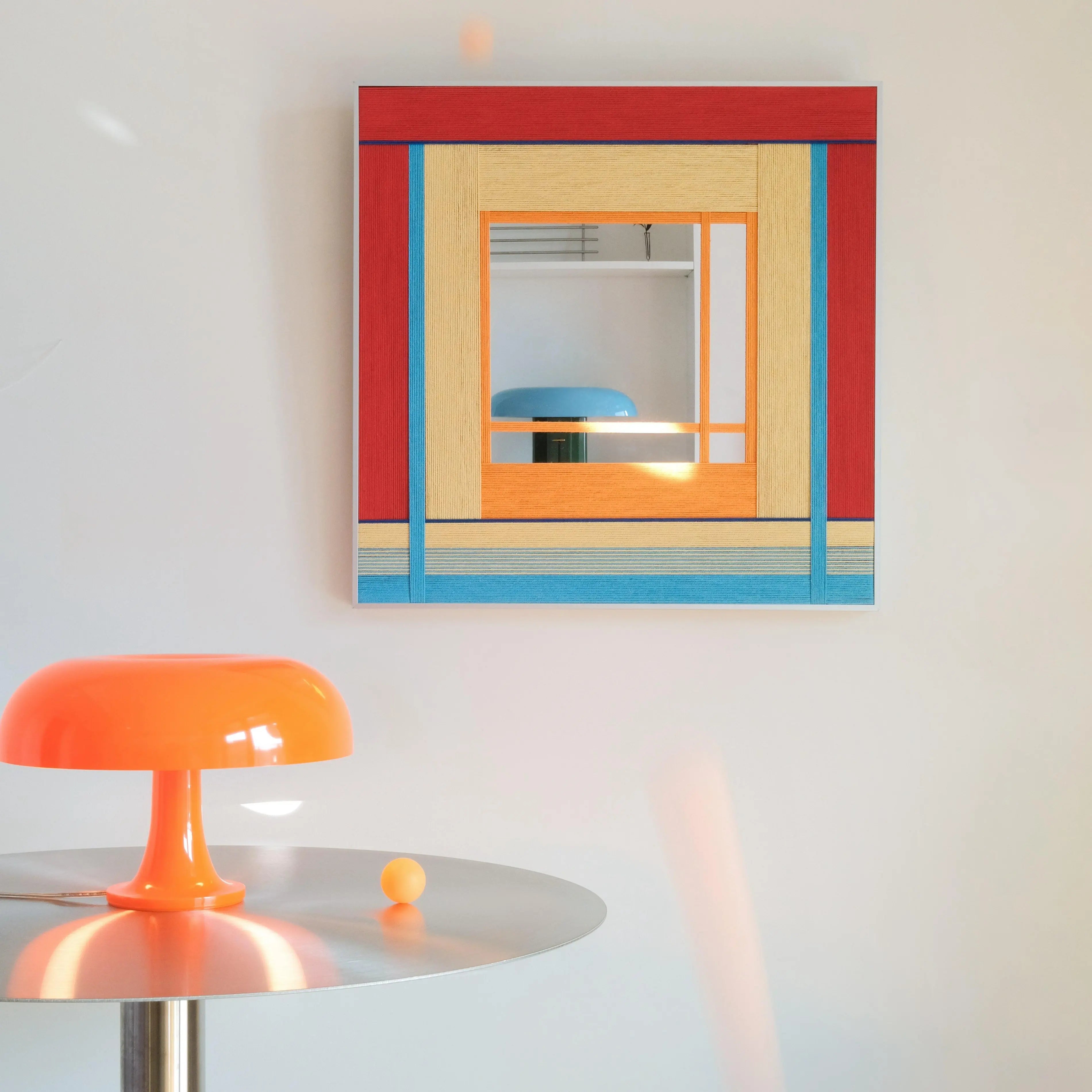 The Fields of Blossoms mirror displayed with a retro orange table lamp, showcasing its bold pop-art inspired geometric color palette.