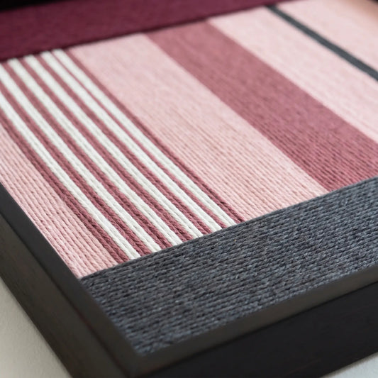 Angled close-up of the Rosy Pulse fiber art showing the dense hand-woven wool texture and sleek black metal frame finish.