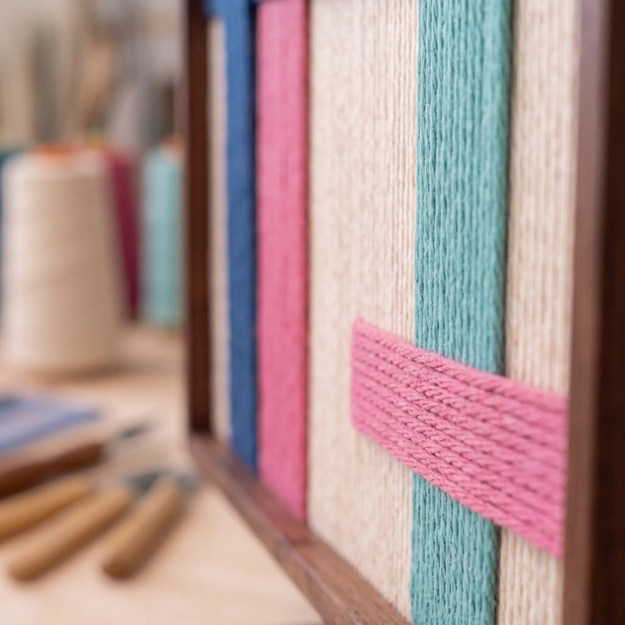 Close-up corner detail of the hand-woven wool tapestry highlighting textured vertical cream and teal yarns with pink bands in a metal frame.