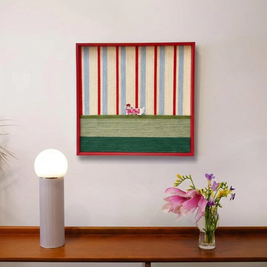 Framed textured fiber wall art styled on a wooden sideboard next to a modern cylindrical lamp and fresh pink flowers.