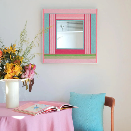 Secret Crush hand‑woven yarn mirror wall art with alternating blush pink, mint green, and soft ivory stripe borders framing a central mirror panel, displayed above a floral vase and pastel decor.
