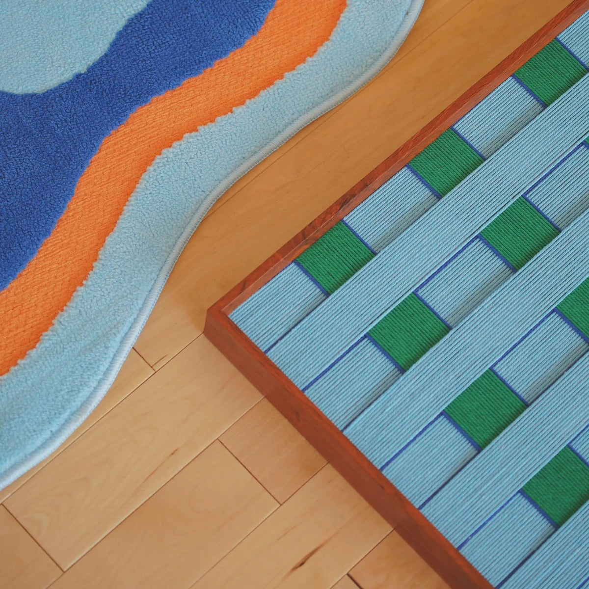 Angled floor detail of Seine Riverbank yarn wall art showing its blue and green wrapped-fiber grid beside a colorful rug.