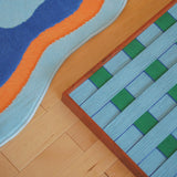 Angled floor detail of Seine Riverbank yarn wall art showing its blue and green wrapped-fiber grid beside a colorful rug.