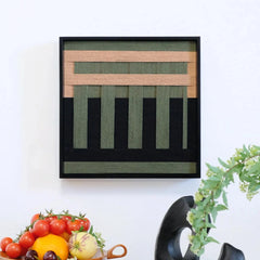 Framed hand-woven wall art featuring dark green and beige geometric blocks on black, styled with fresh fruit and modern decor.
