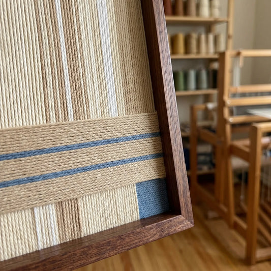 Hand-woven wool tapestry shown from an angled perspective in a sunlit artist studio with a wooden weaving loom in the background.
