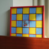 A focused shot of the red-framed soft cotton cord fiber artwork resting on a dark wooden surface, highlighting the high contrast between the frame and woven texture.