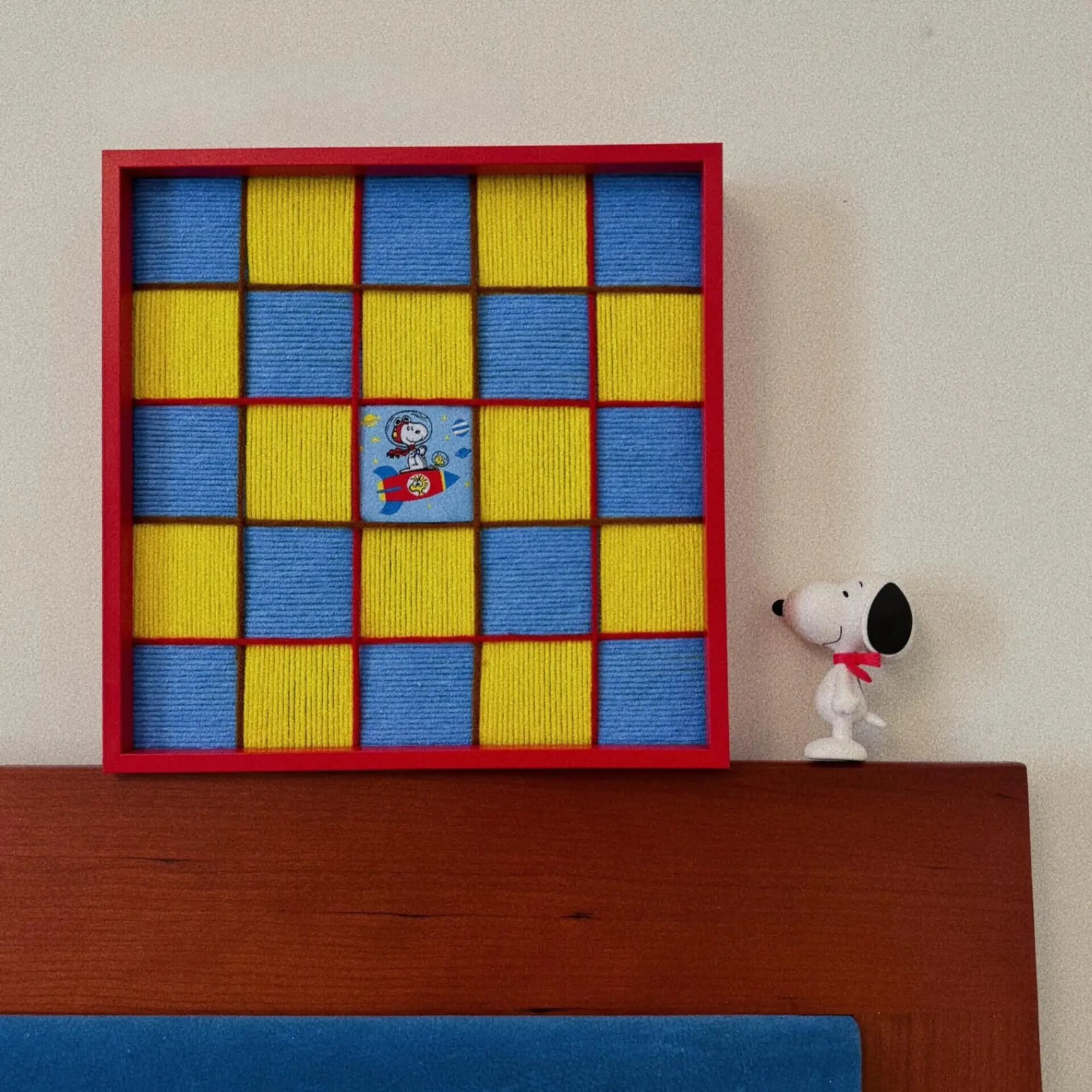 Red framed hand-woven wool wall art featuring a yellow and blue checkerboard pattern with a Snoopy rocket patch, displayed on a wooden headboard.