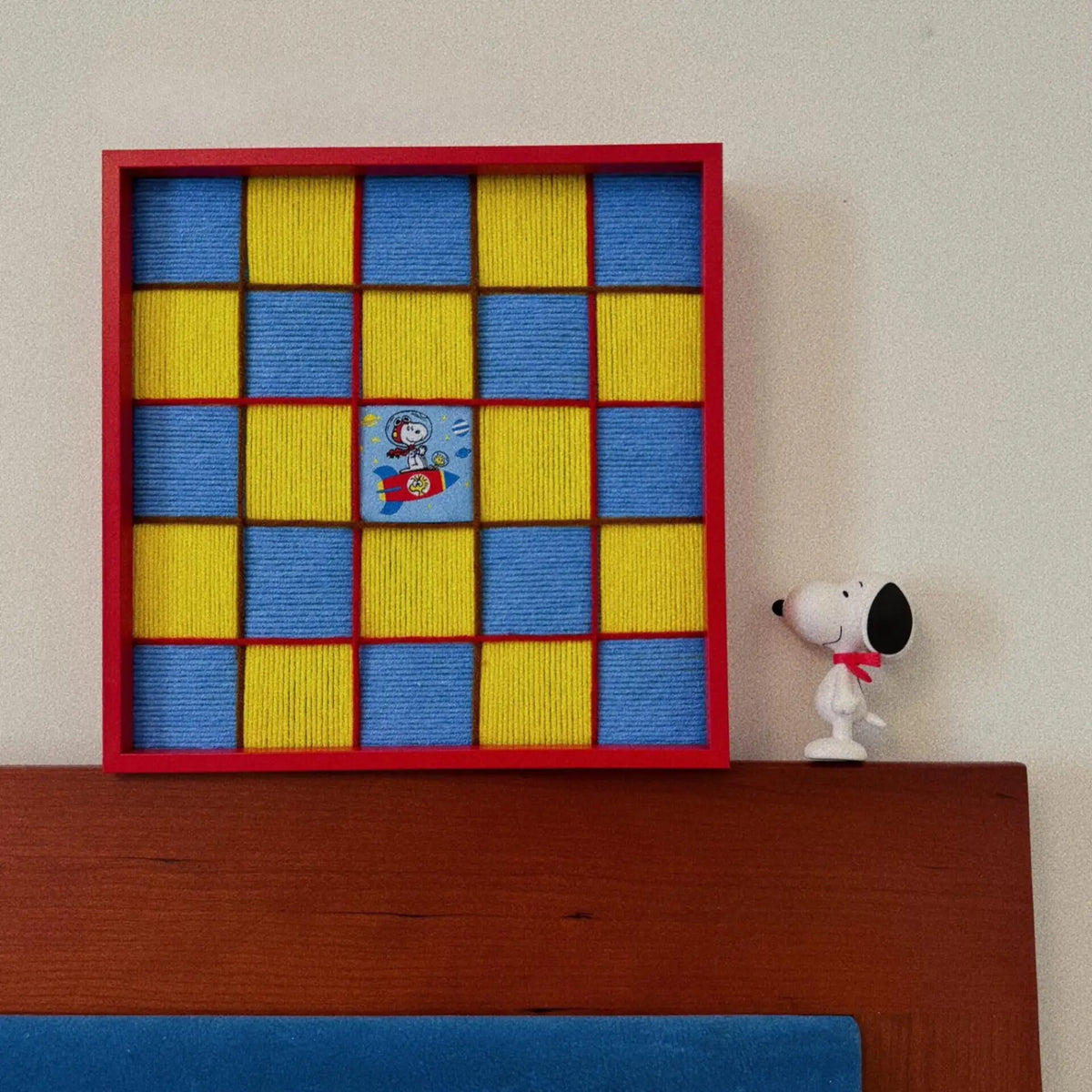 Red framed soft cotton cord wall art featuring a yellow and blue checkerboard pattern with a Snoopy rocket patch, displayed on a wooden headboard.