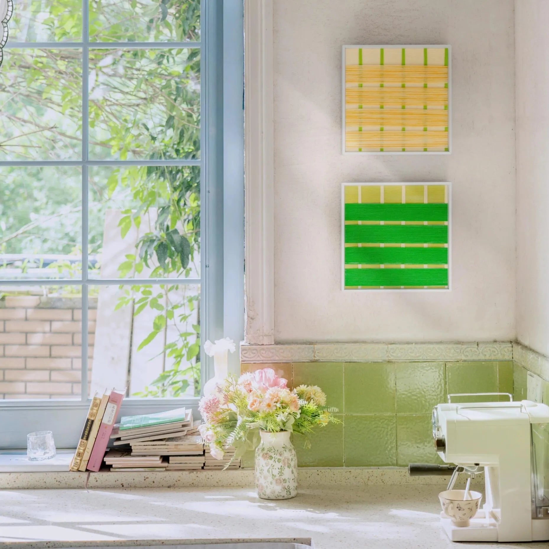 Two framed hand-woven wool art pieces hanging vertically near a sunny window, featuring fresh green and yellow spring-inspired patterns.