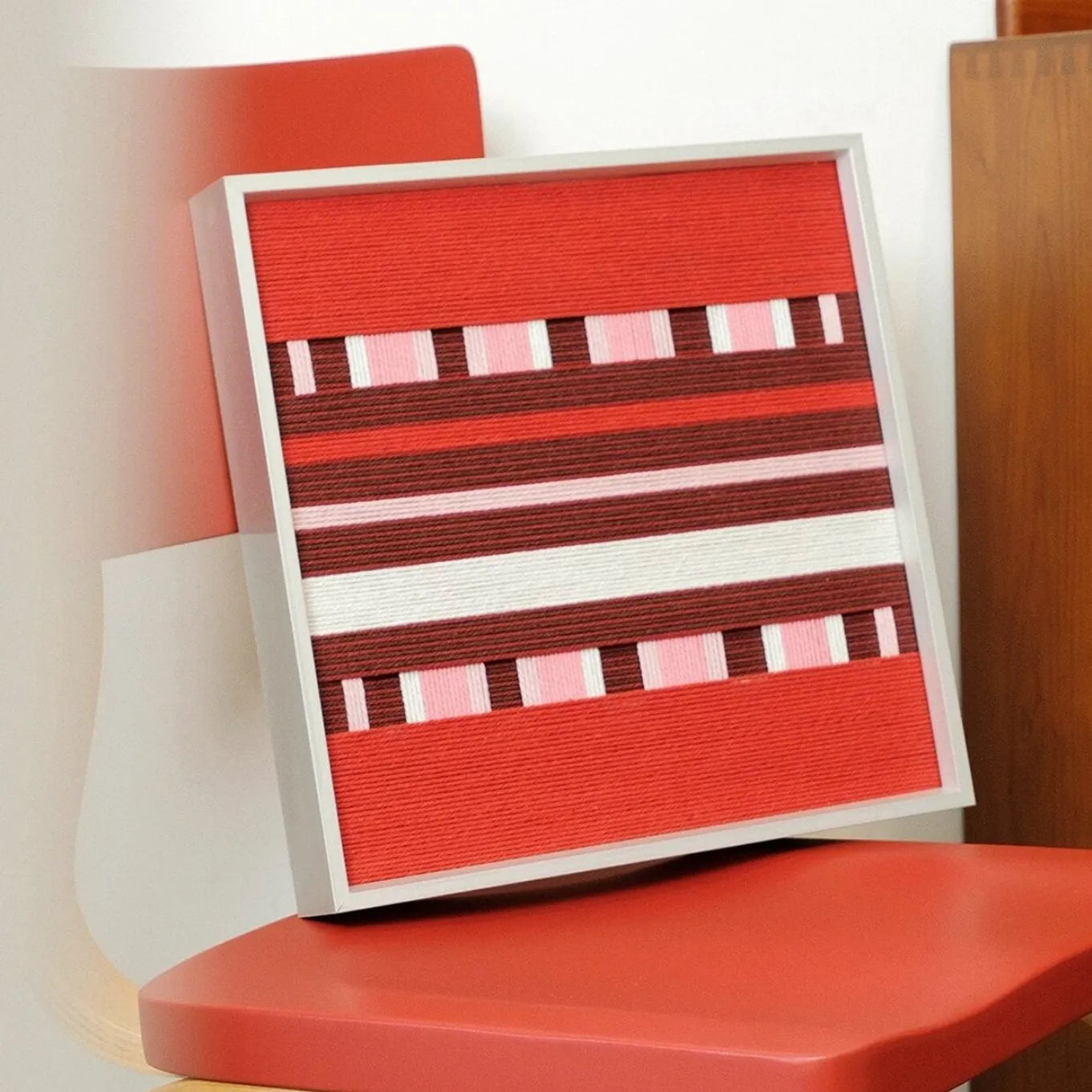 Angled view of Spring Miss yarn wall art leaning on a red chair, highlighting the textured wool weave and bold stripe pattern.
