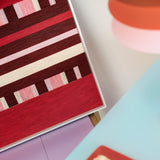 Angled close-up of Spring Miss SQ029 showing red burgundy pink and cream yarn bands and the white frame