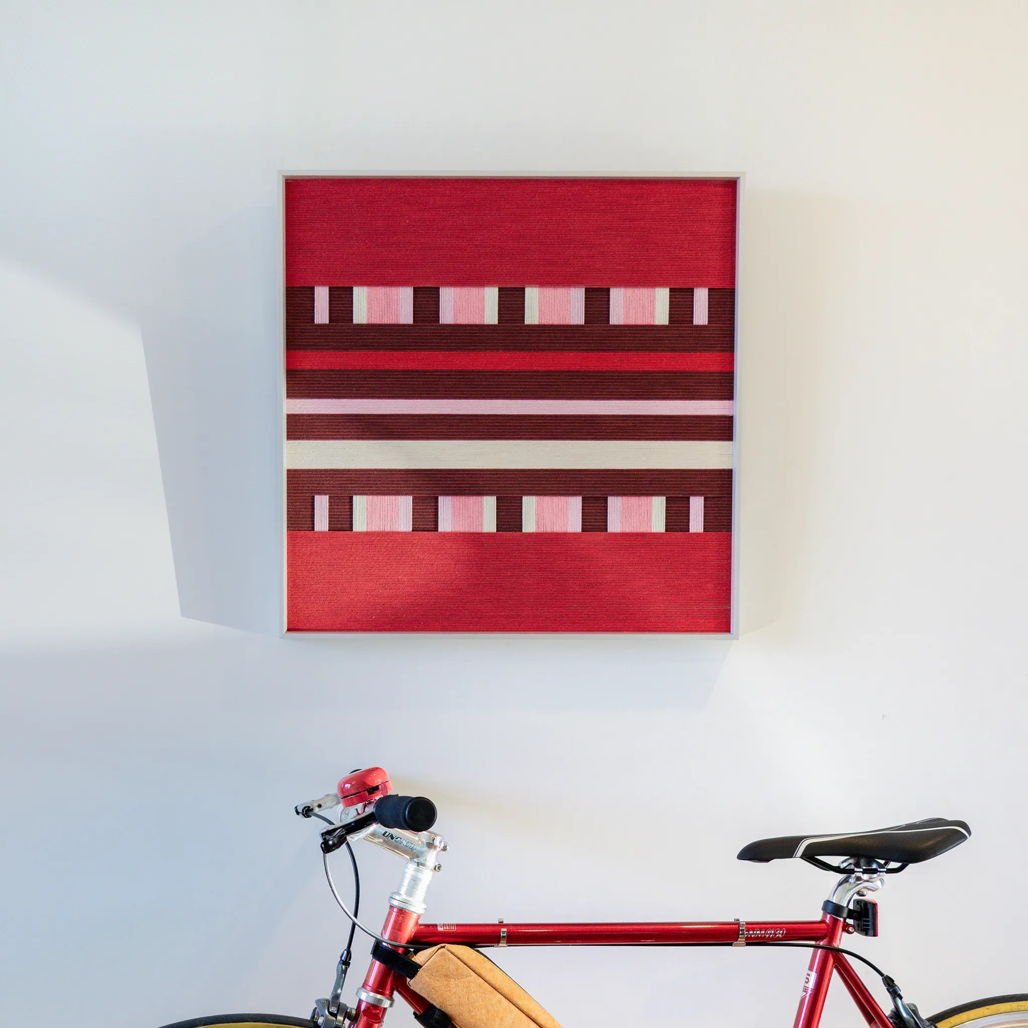 Spring Miss SQ029 red and burgundy framed yarn art displayed on a white wall above a red bicycle
