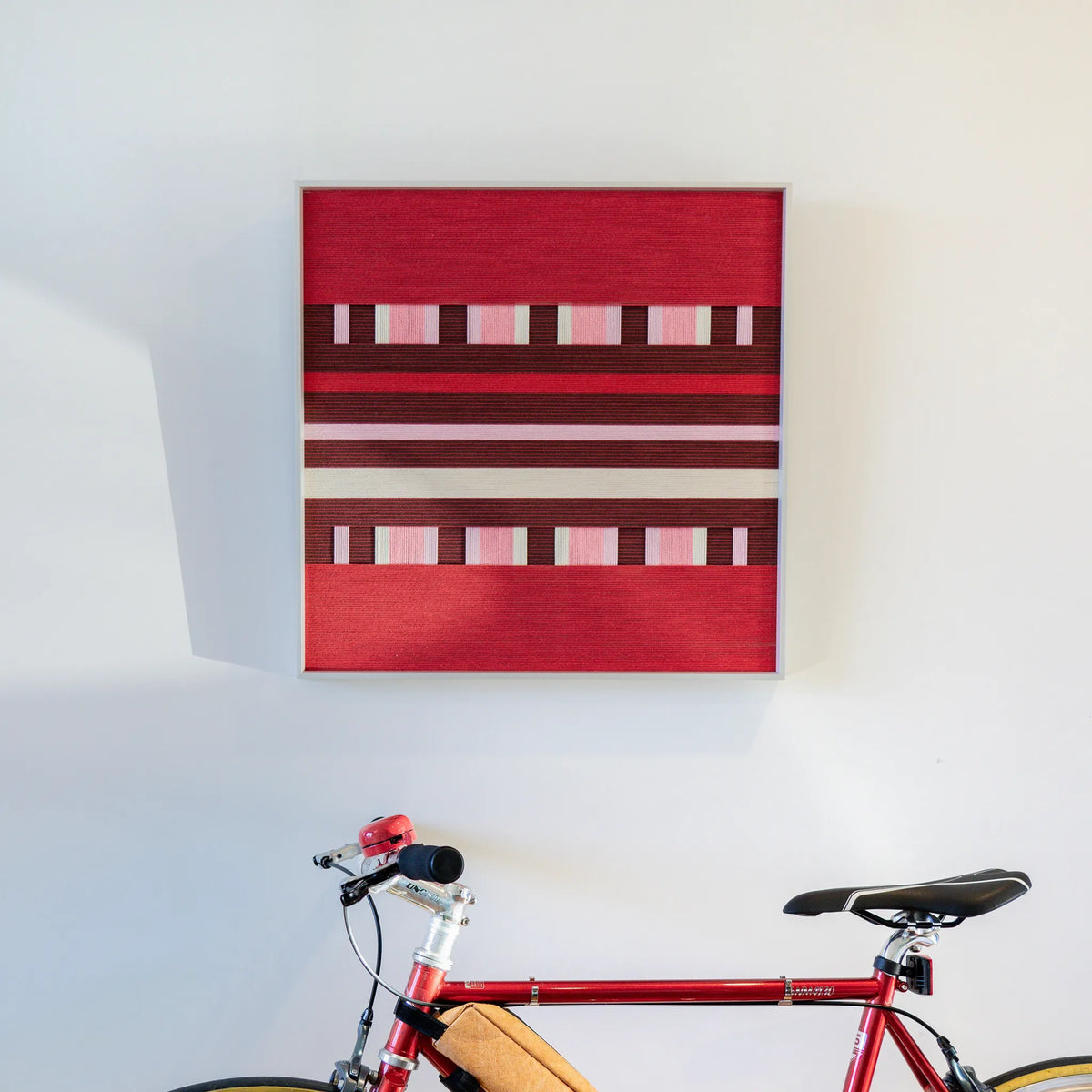Spring Miss SQ029 red and burgundy framed yarn art displayed on a white wall above a red bicycle
