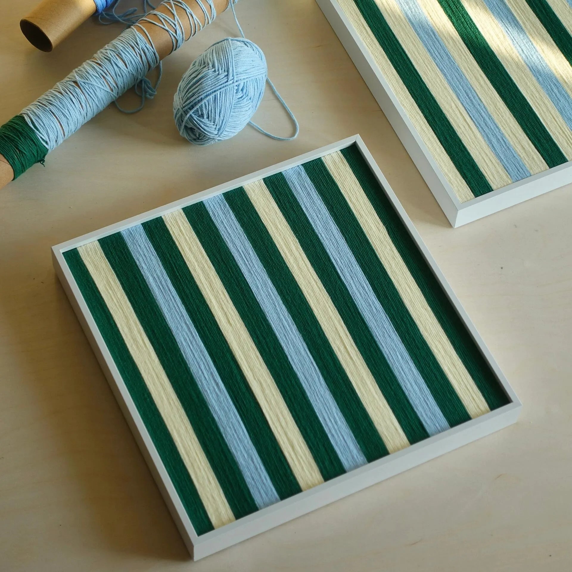 Single Spring Lake SQ039 green blue and cream striped framed yarn artwork lying on a studio worktable beside yarn materials