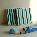 Two Spring Lake SQ039 green blue and cream striped framed yarn artworks displayed with yarn balls and wrapped spools in the studio