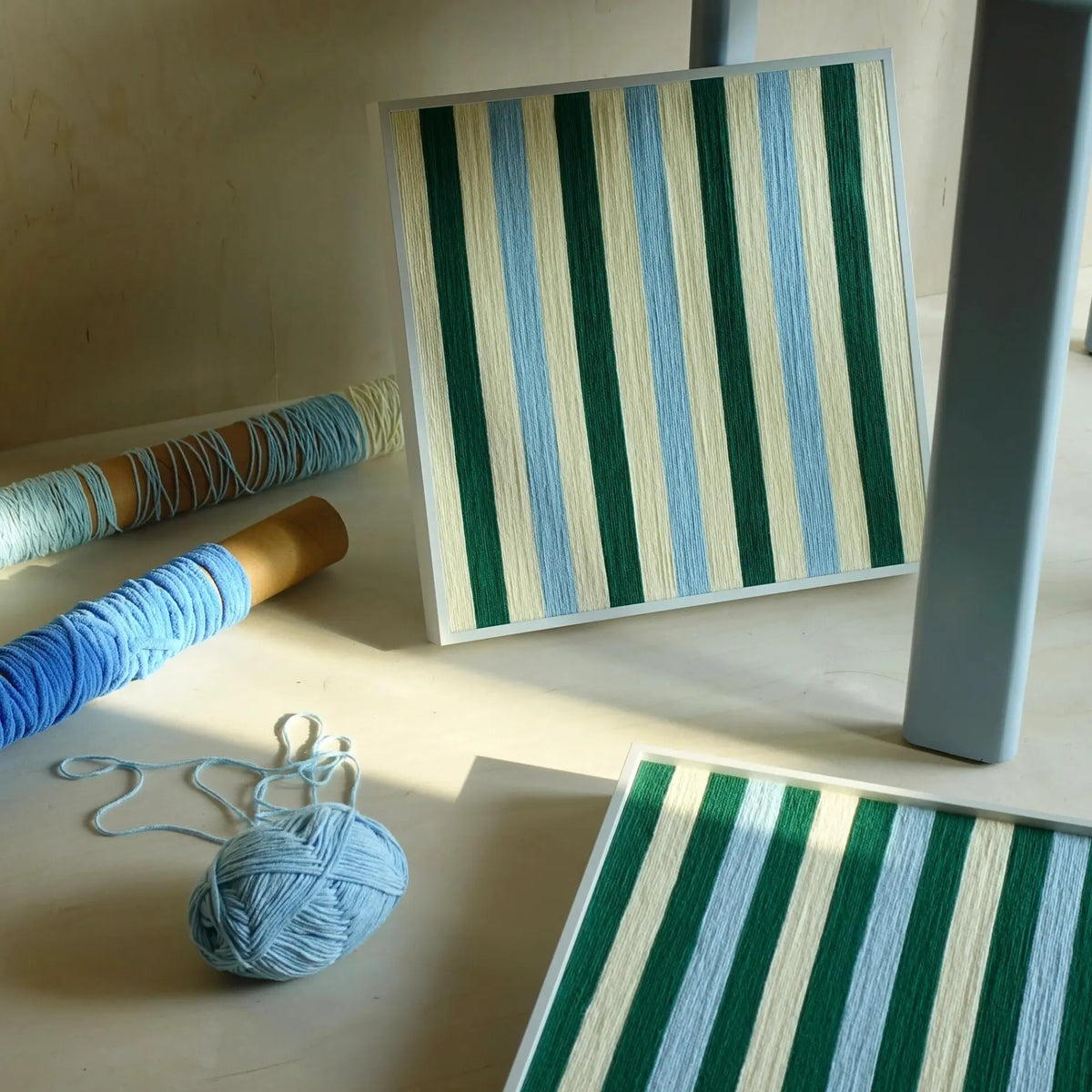 Spring Lake SQ039 green blue and cream striped framed yarn artwork standing upright in sunlight beside yarn balls and wrapped spools