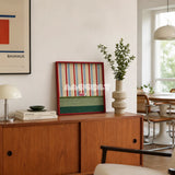 Scarlet Stripe Pup Saunter SQ045 striped framed yarn art with a dog motif displayed on a wood sideboard beside a Bauhaus poster and plant