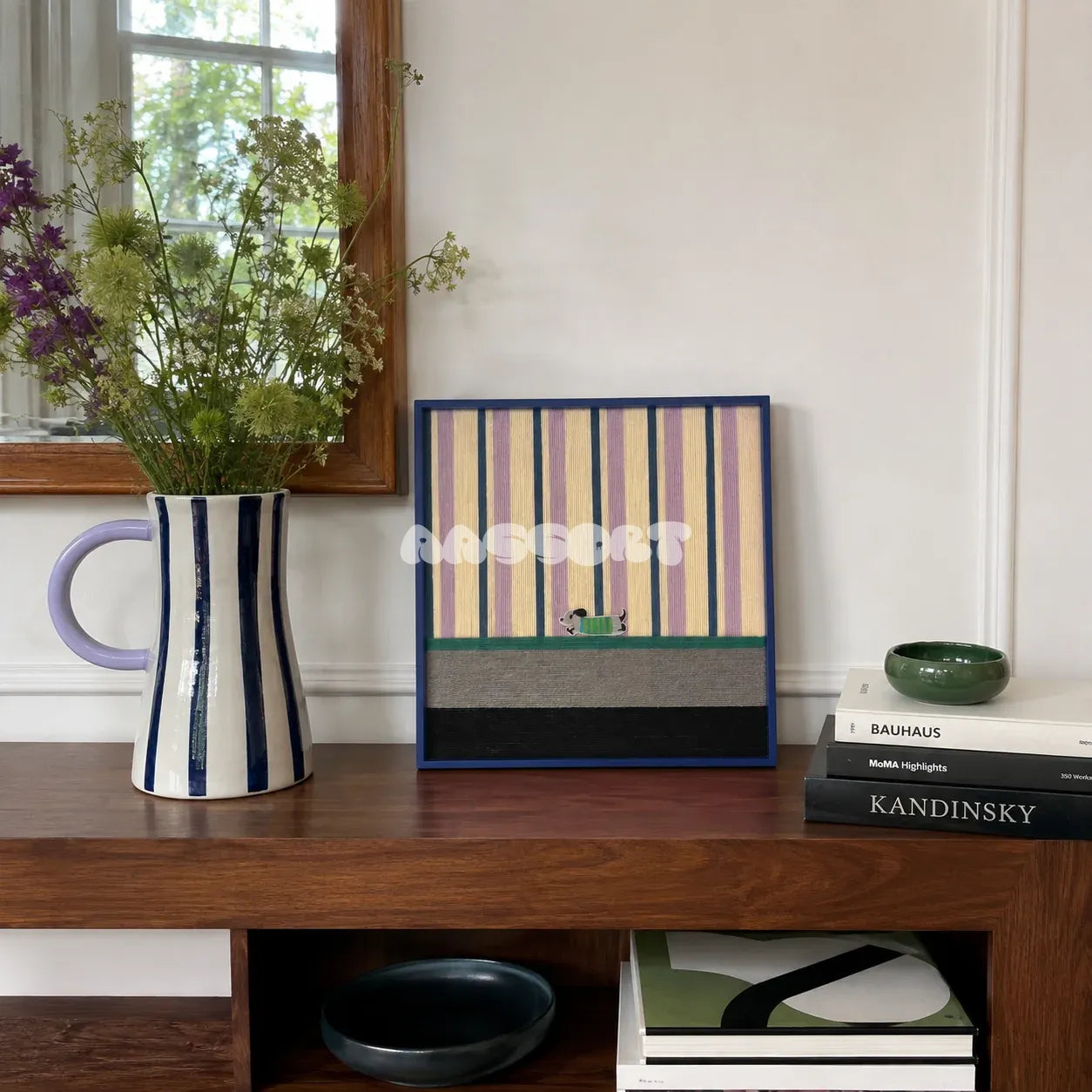 Cerulean Stripe Pup Frolic SQ047 striped framed yarn art with a dog motif displayed on a wood sideboard beside flowers and art books