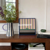 Cerulean Stripe Pup Frolic SQ047 striped framed yarn art with a dog motif displayed on a wood sideboard beside flowers and art books