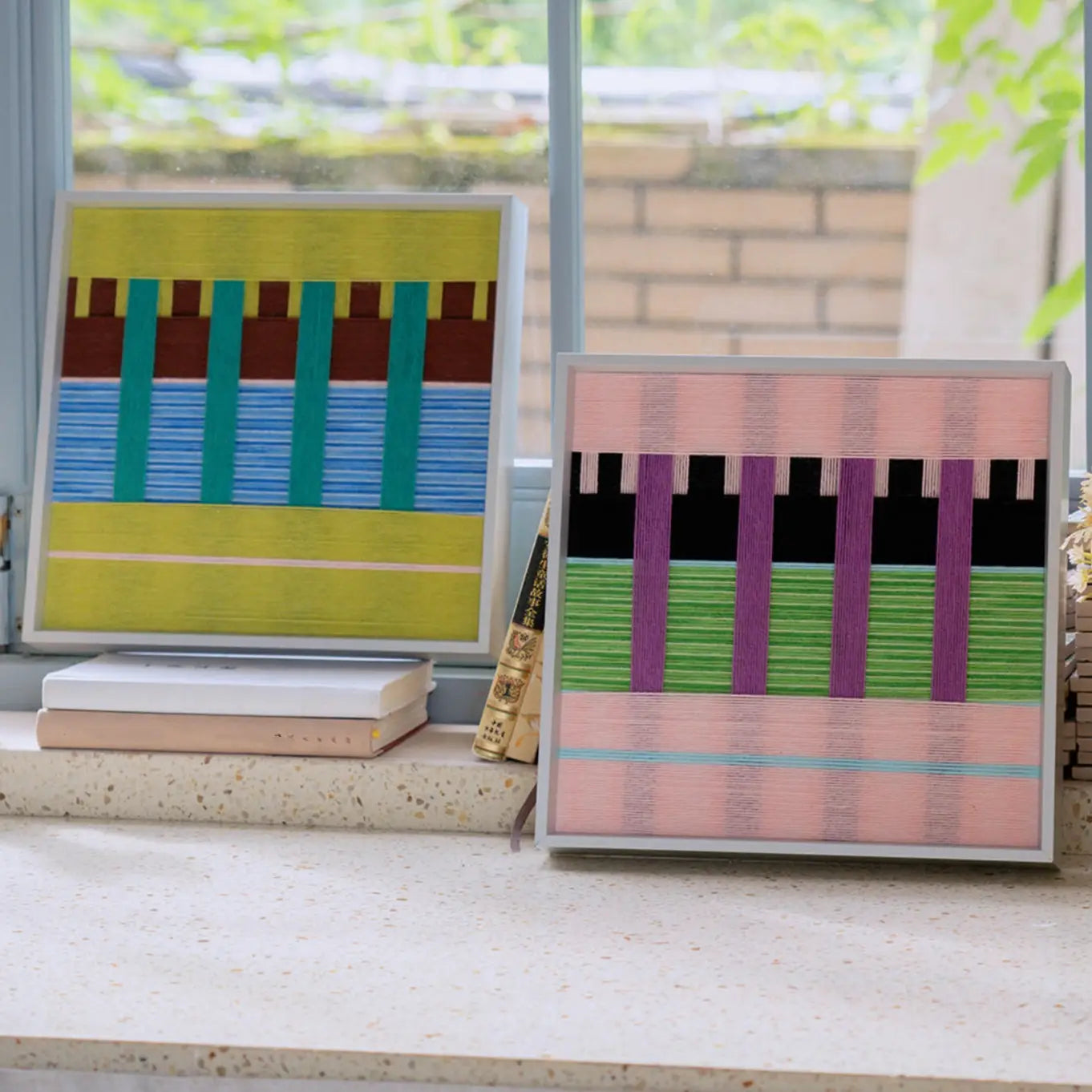 Woodland Mirror and Pastel Nocturne SQ116 textile artworks displayed together on stacked books near a bright window.