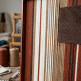 Angled close-up of Mocha Prelude SQ124 showing the frame and dimensional mocha block over earth-tone yarn stripes.