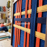 Detail of Silent Axis framed textile wall art, featuring navy, terracotta, and wheat yellow cotton cord arranged in an architectural grid.
