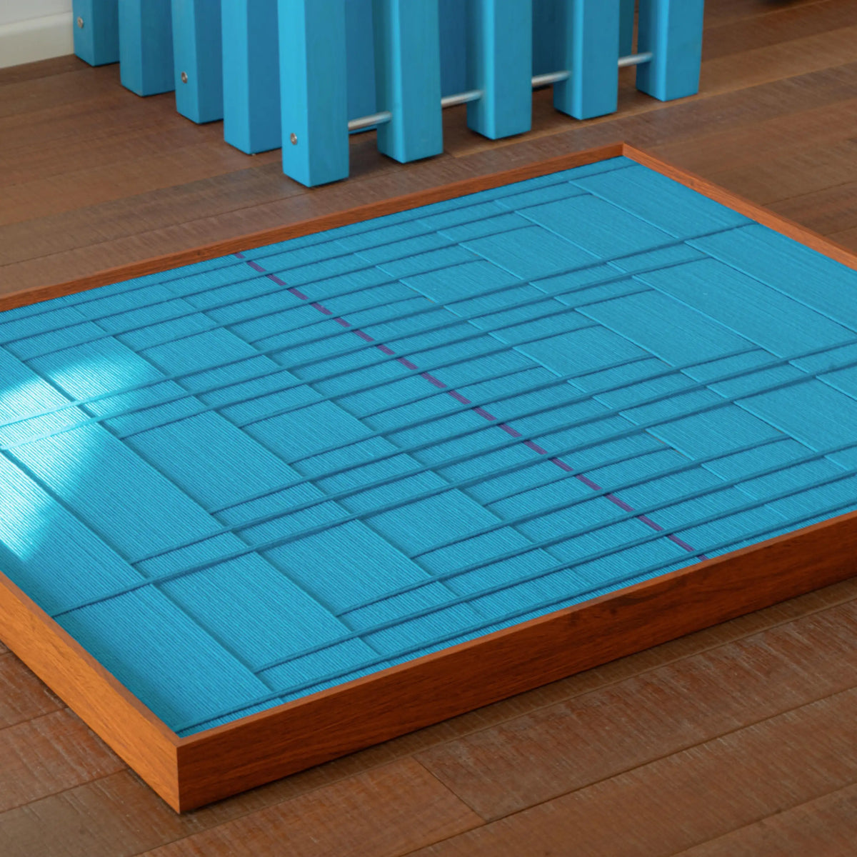 High-angle view of the blue geometric fiber art lying flat on a wooden floor, emphasizing the depth and structure of the woven pattern.