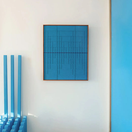 Framed "Submersion" hand-woven wall art featuring a monochromatic blue geometric design, styled on a white wall with vertical blue accents.
