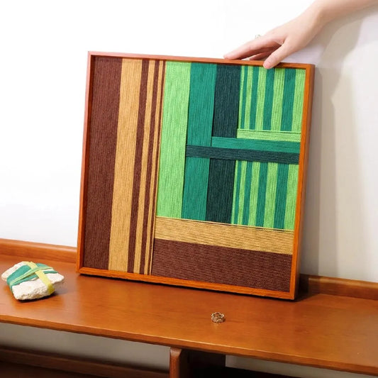 Summer Mountain Walk hand-woven wool yarn wall art featuring alternating forest green, olive, and chocolate brown stripe panels in a natural wood frame, displayed on a console table.
