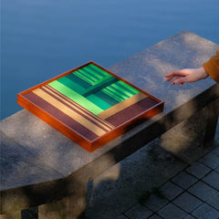 Summer Mountain Walk wall art placed beside a mountain lake, emphasizing the contrast between vibrant green yarn stripes and natural setting.
