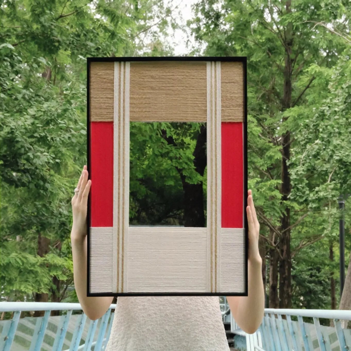 Hand-held view of The Sun Always Rises yarn mirror art against a lush green background, showing its compact square form and vibrant stripe contrast.
