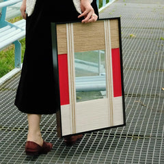 Hand-held view of The Sun Always Rises yarn mirror art being carried along a metal walkway, emphasizing its sturdy frame and crisp red accent stripe against neutral wool panels.
