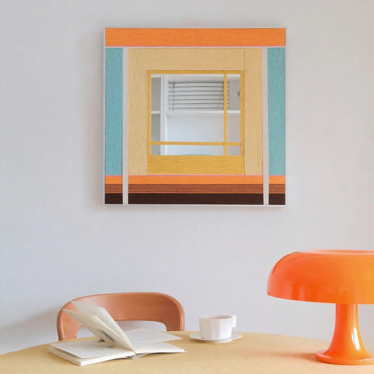 Sun‑Drenched Days hand‑woven yarn wall art with pastel peach, mustard yellow, and sky‑blue vertical stripes in a natural wood frame, displayed above a desk with an orange lamp.
