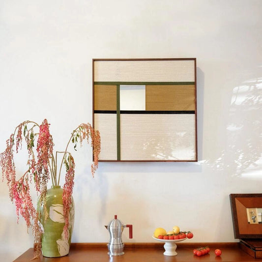 Through the Mirror hand‑woven wool yarn mirror wall art featuring alternating taupe, olive green, and cream stripe panels framing a central reflective panel, displayed above a tabletop with vase and coffee pot.
