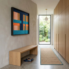 Vibrant square fiber art hanging in a bright entryway above a light wood minimalist bench and jute runner rug.