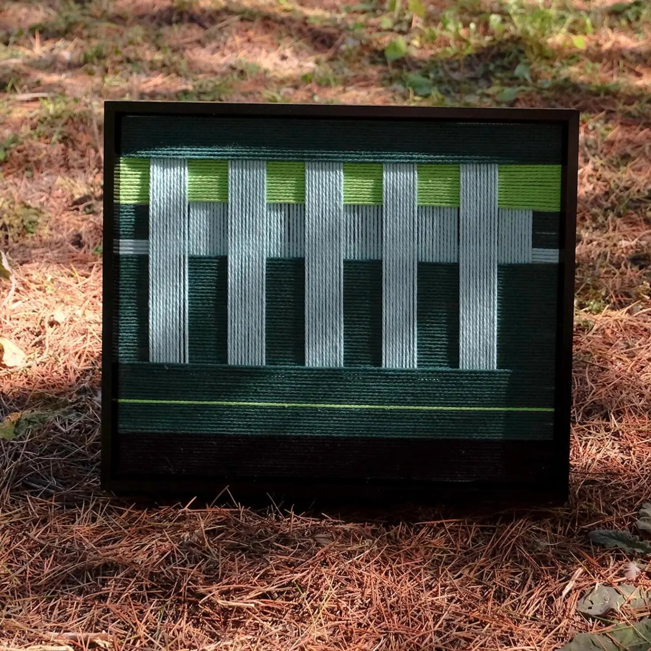 Velvet Vine hand-woven wall art resting on forest floor, showcasing its square format and alternating green-white stripes in a minimalist frame
