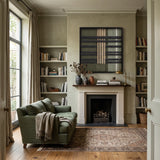 The Verdant Silence hand-woven wool tapestry hangs above a classic fireplace in a cozy living room with olive green furniture.
