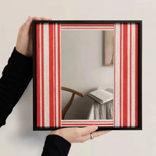 Wave Tide hand-woven yarn mirror wall art with central reflective panel framed by alternating red and white wool stripe borders in a sleek black wood frame, displayed above a chair and open book.
