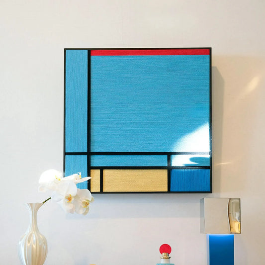 Weave Reshape hand-woven wool yarn wall art featuring sky-blue panel, mustard-yellow stripe, red base block, and black accent band in a natural wood frame, displayed above a console table.
