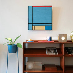 Weave Reshape yarn wall art styled on a desk with a white lamp and decorative orb, showcasing its precise color-block grid and sturdy frame.
