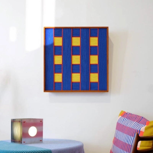 Window Peek at Moon hand-woven yarn art mounted on a console table with lamp and ceramic jar, showcasing its stair-step blue gradient design and natural wood frame
