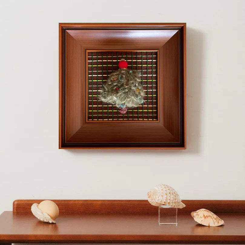 The framed wool tapestry hanging above a simple wooden shelf, adding a touch of handcrafted texture to a clean wall space.