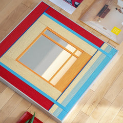 Flat lay view of the textured fiber mirror on a wooden floor, highlighting the vibrant red, blue, and yellow wool frame construction.